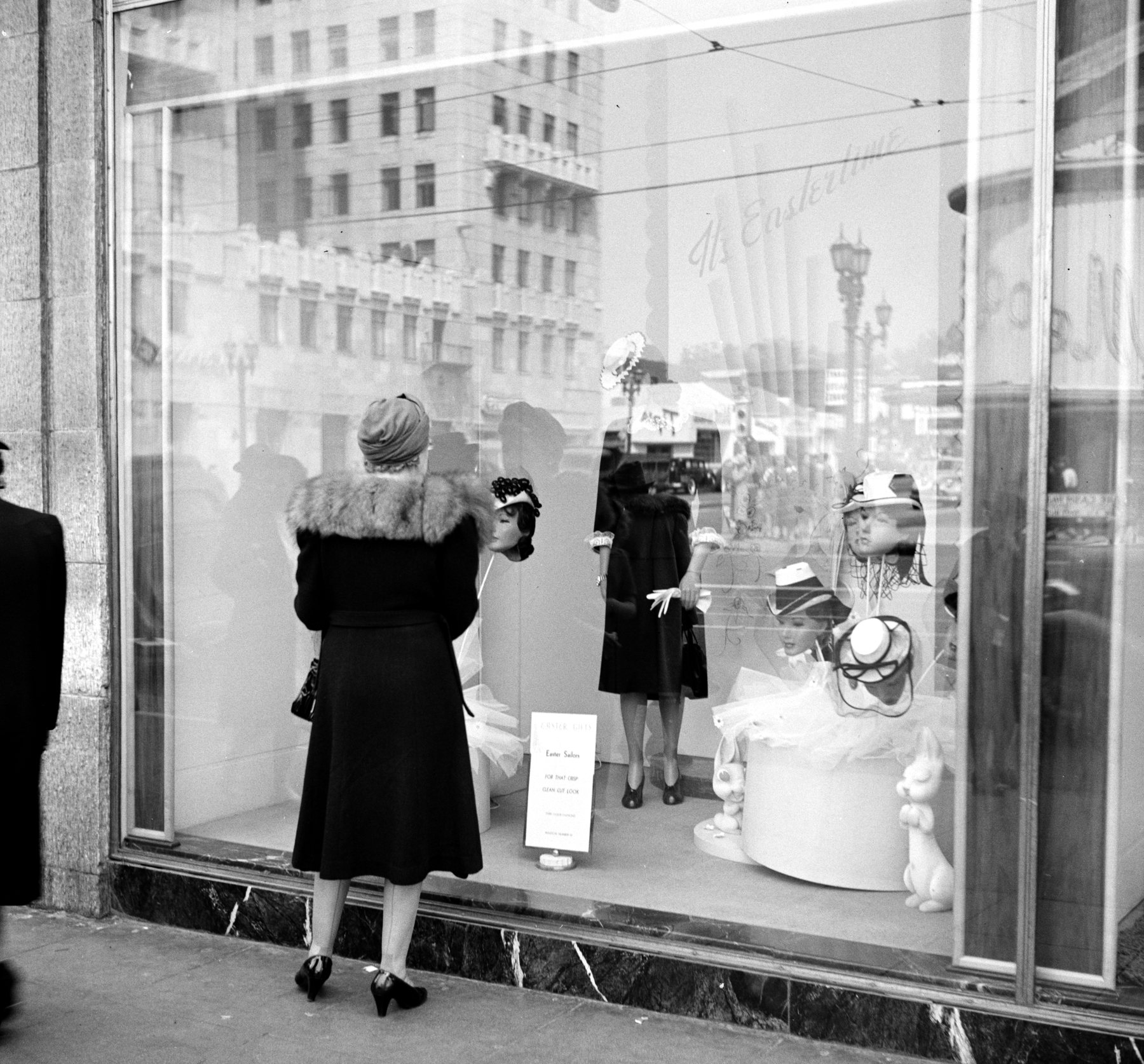 A woman in a fur-collared coat, seen from behind, studying the hats in a department store window.