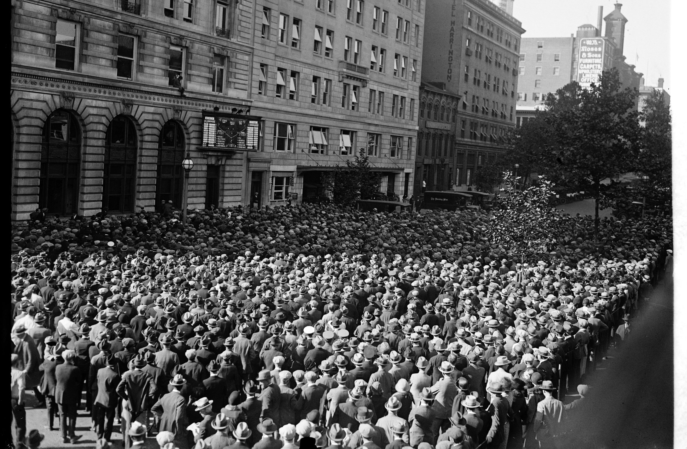 A packed street crowd facing a public scoreboard.