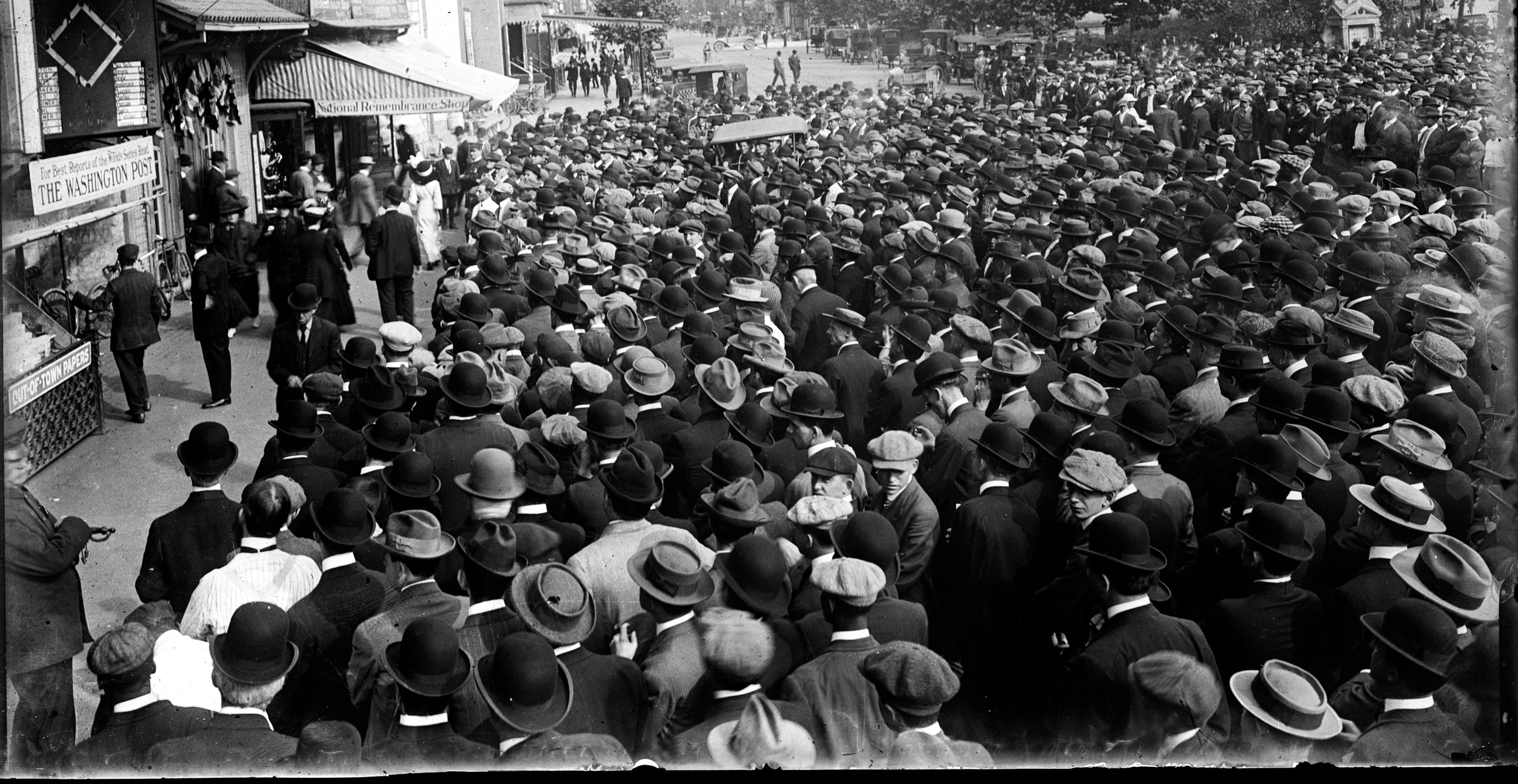 A street packed with men in hats, seen from behind, waiting at a scoreboard.