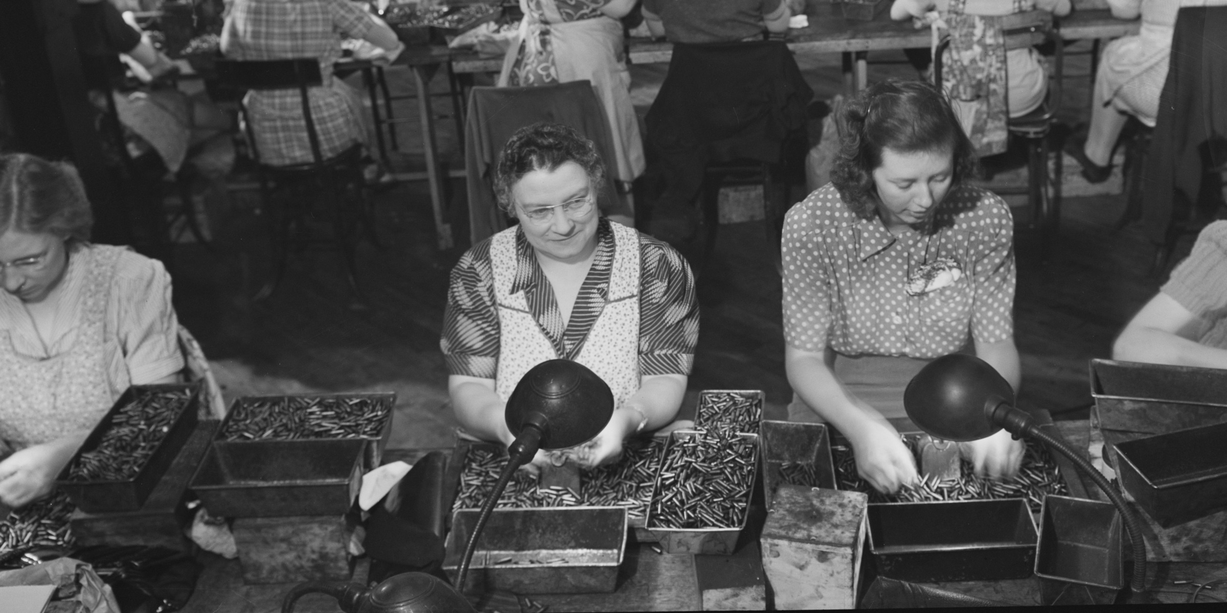 Women gauge inspectors at a long factory bench, checking small parts from metal trays under desk lamps.